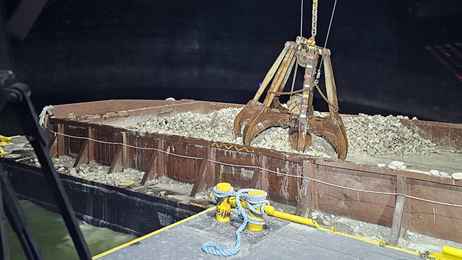 A nighttime photo of a grapple grabbing a pile of cobble from a barge. The scene is lit with work lights. A large cleat is in the foreground of the photo on the edge of the barge the photographer is standing on.